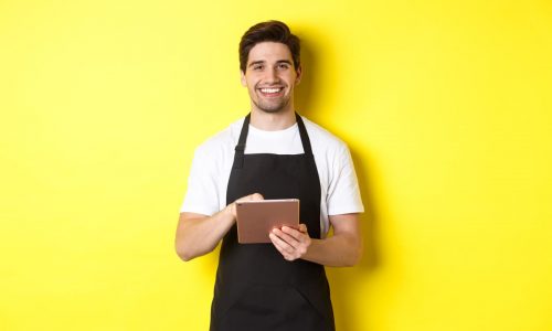 guapo-camarero-tomando-ordenes-sosteniendo-tableta-digital-sonriendo-vestido-uniforme-delantal-negro-pie-sobre-fondo-amarillo (1)